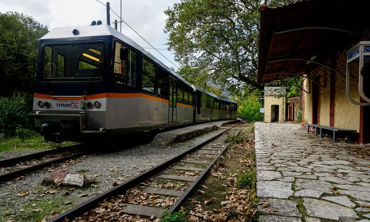 Ein Zug nach Griechenland steht auf den Gleisen neben einem alten Bahnhofsgebäude. Herbstlaub bedeckt den Bahnsteig.