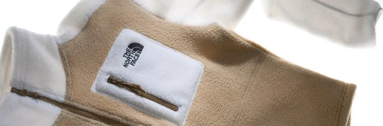 Ein beige-weißes Fleece-Oberteil von The North Face mit Reißverschlusstasche. Fleece ist ein weiches, warmes Material.