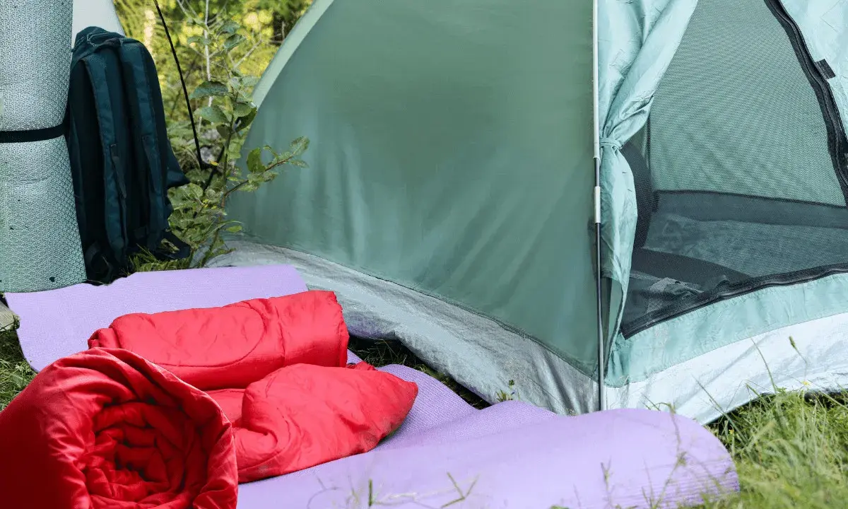 Zwei rote Schlafsäcke liegen auf lila Isomatten vor einem grünen Zelt. Ein Rucksack und eine weitere Isomatte stehen daneben.