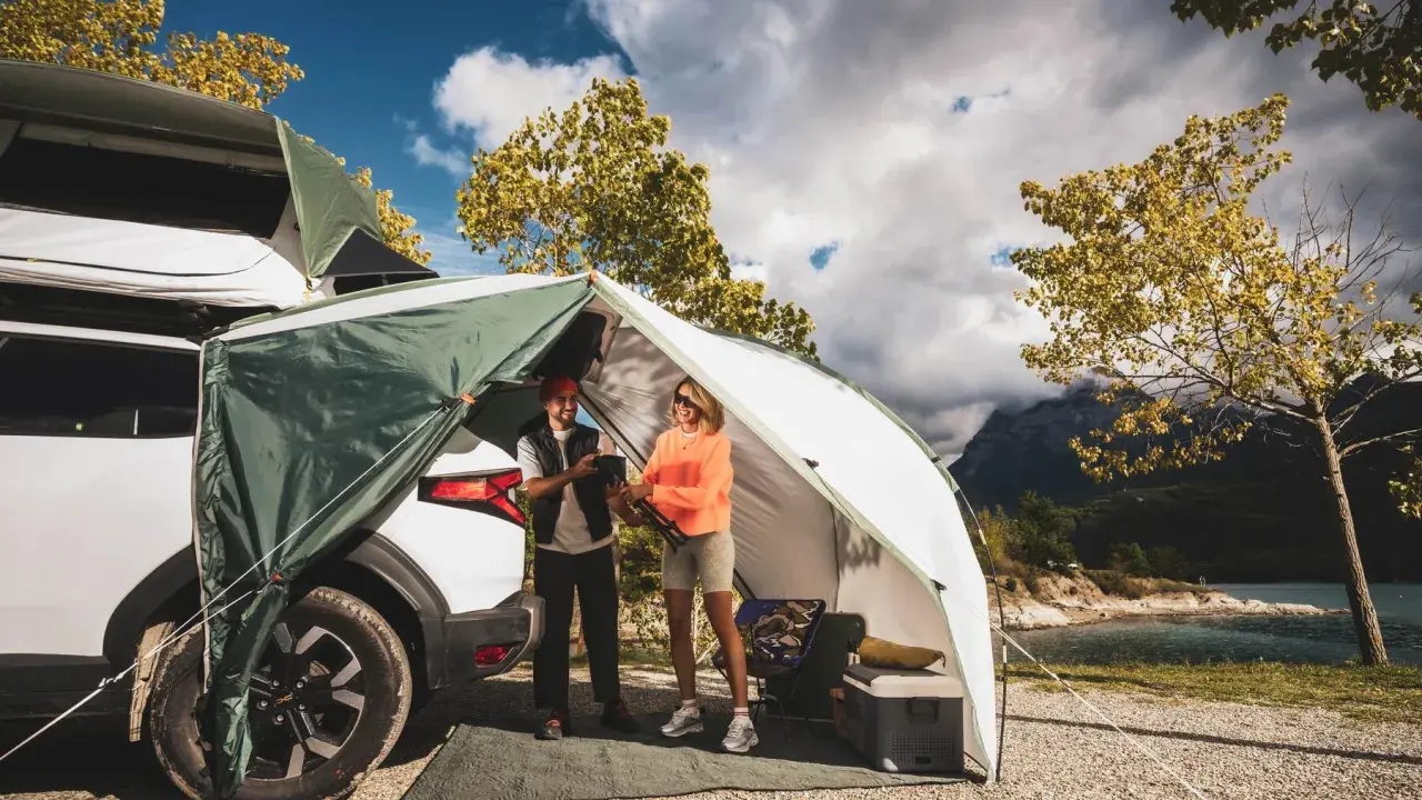 Zwei Frauen genießen das Frauen Camping Abenteuer am See. Ein Zelt ist am Auto befestigt, mit Blick auf Berge und Wasser.