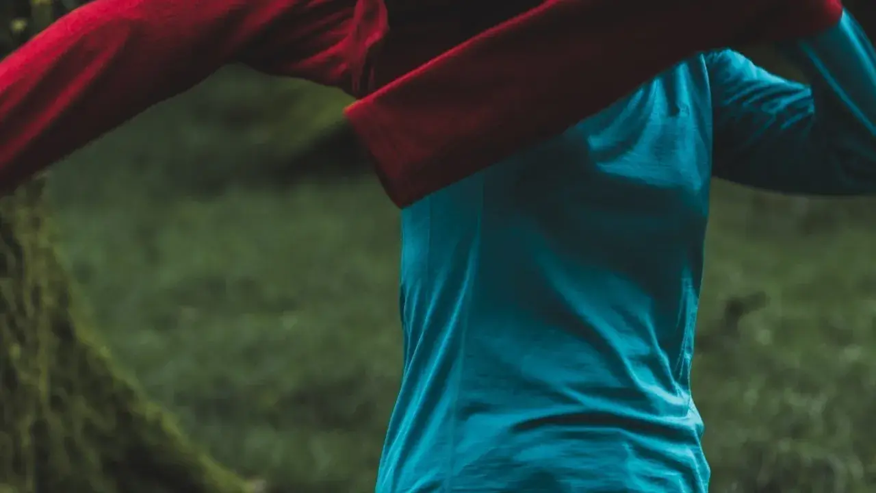Person zieht ein rotes Oberteil über ein blaues Funktionsshirt. Das blaue Shirt ist ein Baselayer, der die Körperwärme reguliert.