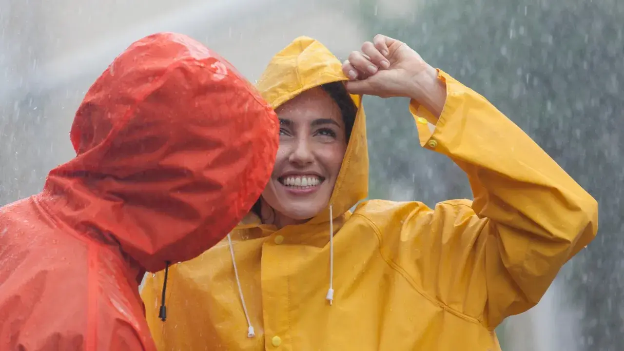 Zwei lachende Personen im Regen, eine in Gelb, die andere in Rot. Die gelbe Regenjacke ist im Fokus.