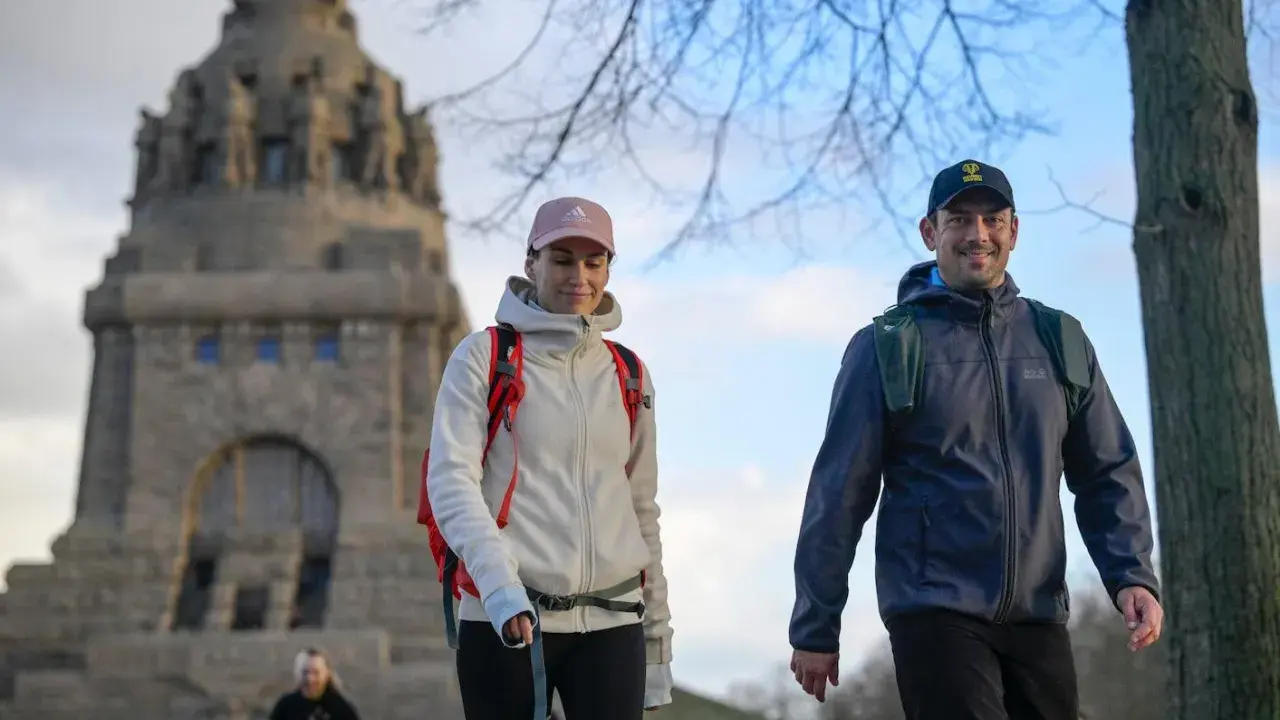 Ein Paar genießt eine Wanderung, passend mit wetterfester Wanderkleidung und Rucksäcken ausgestattet, vor einem imposanten Denkmal.