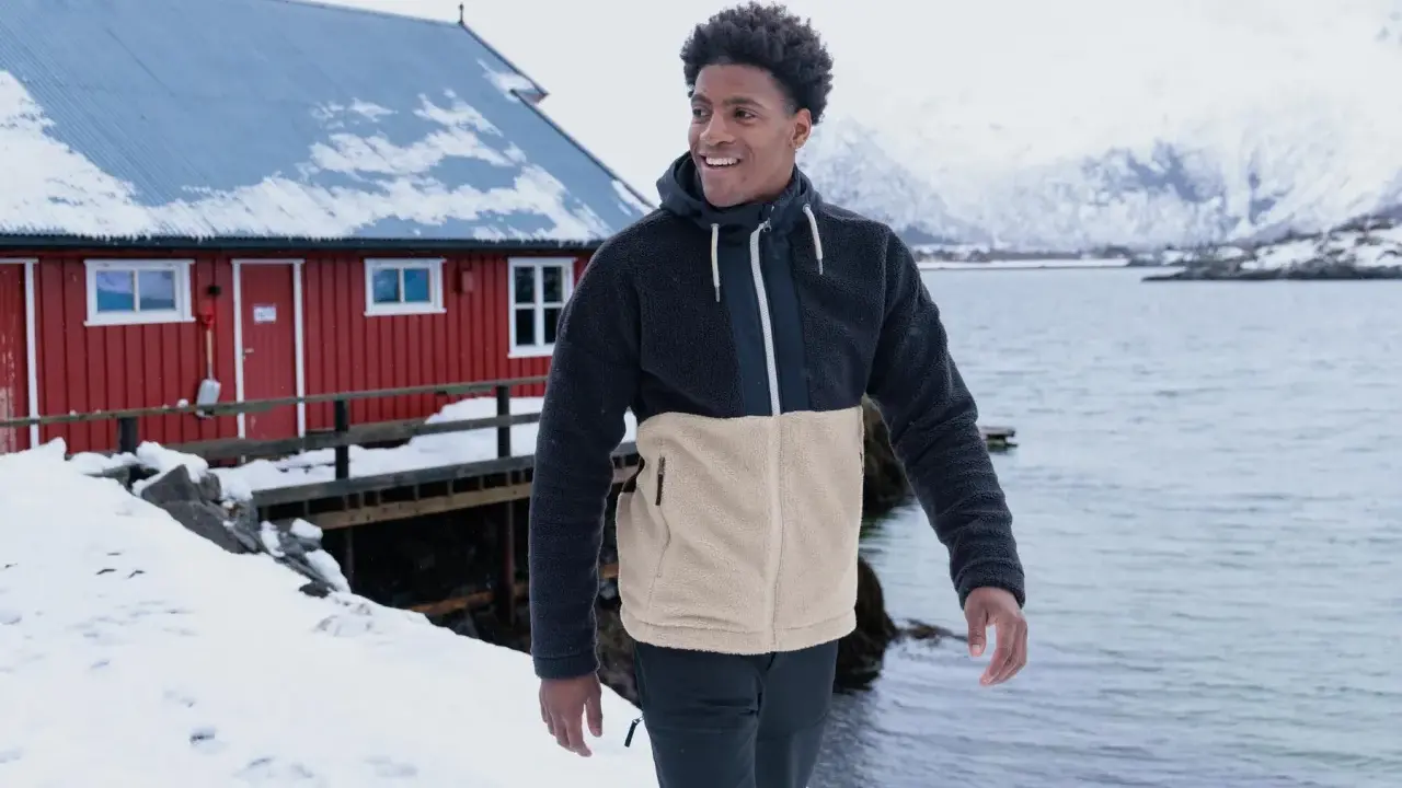 Mann in warmer Fleecejacke, bereit für das Waschen, steht vor einem roten Haus am Meer.
