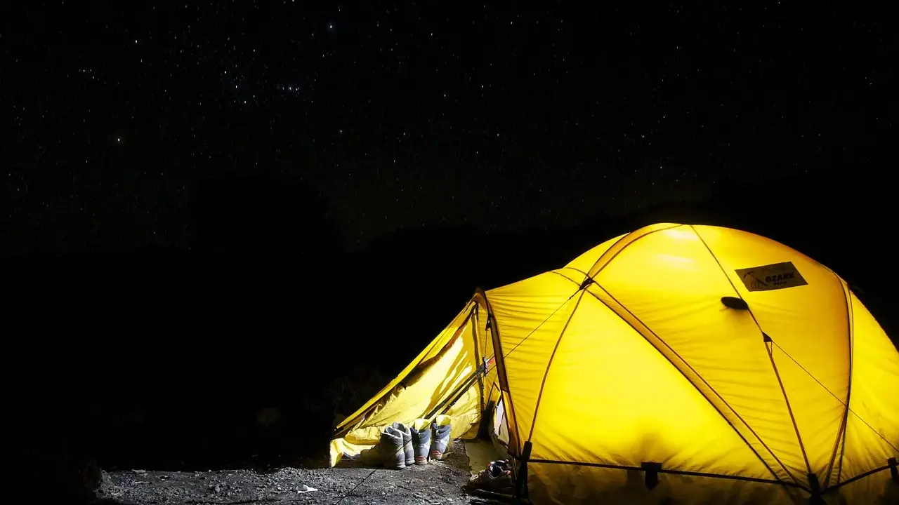 Gelbes Zelt leuchtet in der Nacht unter Sternenhimmel. Stiefel stehen bereit. Alles, was man zum Zelten braucht, ist hier.