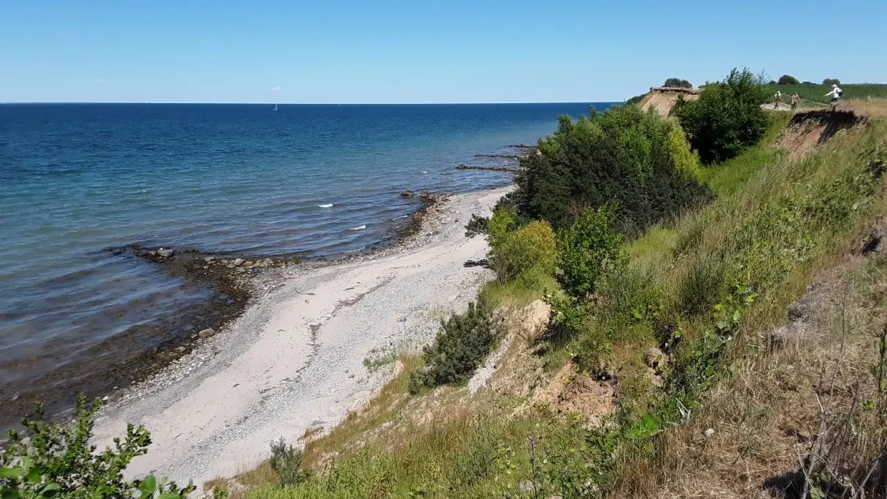 Ein sonniger Tag am Timmendorfer Strand. Die Ostsee glitzert, ein schmaler Kiesstrand lädt zum Spaziergang ein, während grüne Büsche die Steilküste säumen.