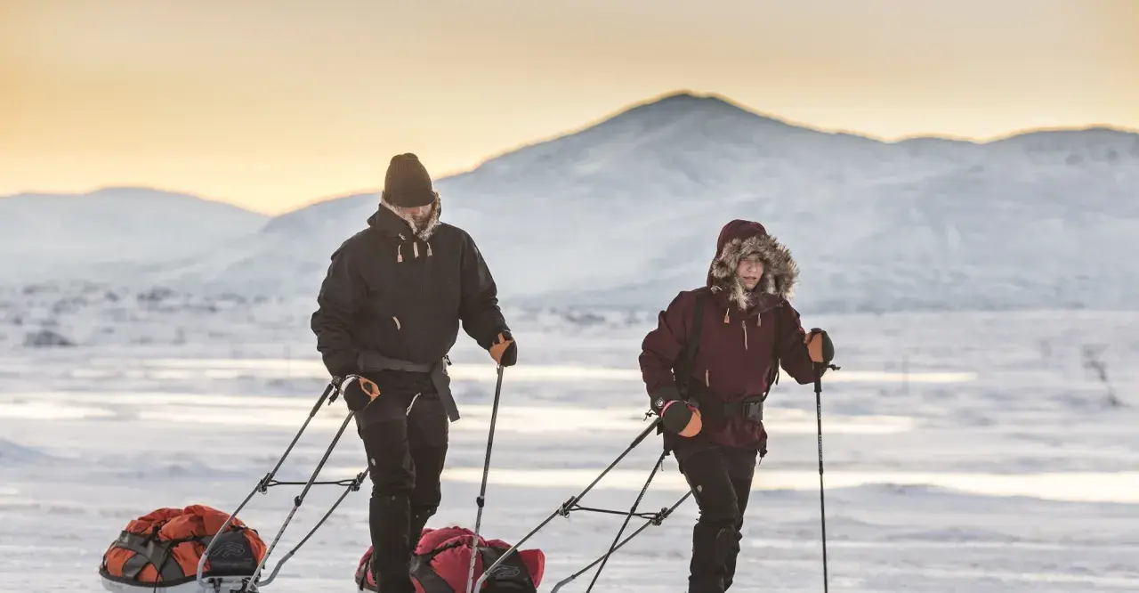 Zwei Personen ziehen Schlitten durch verschneite Landschaft. Die Berge im Hintergrund sind mit Schnee bedeckt. Ein Abenteuer wie aus dem Fjällräven g-1000 Katalog.