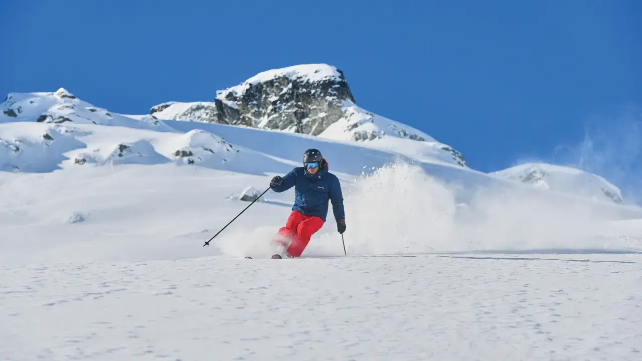 Skifahrer in roter Hose und blauer Jacke wirbelt Schnee auf. Die Columbia Omni-Heat Erfahrung: warm und trocken im Pulverschnee.
