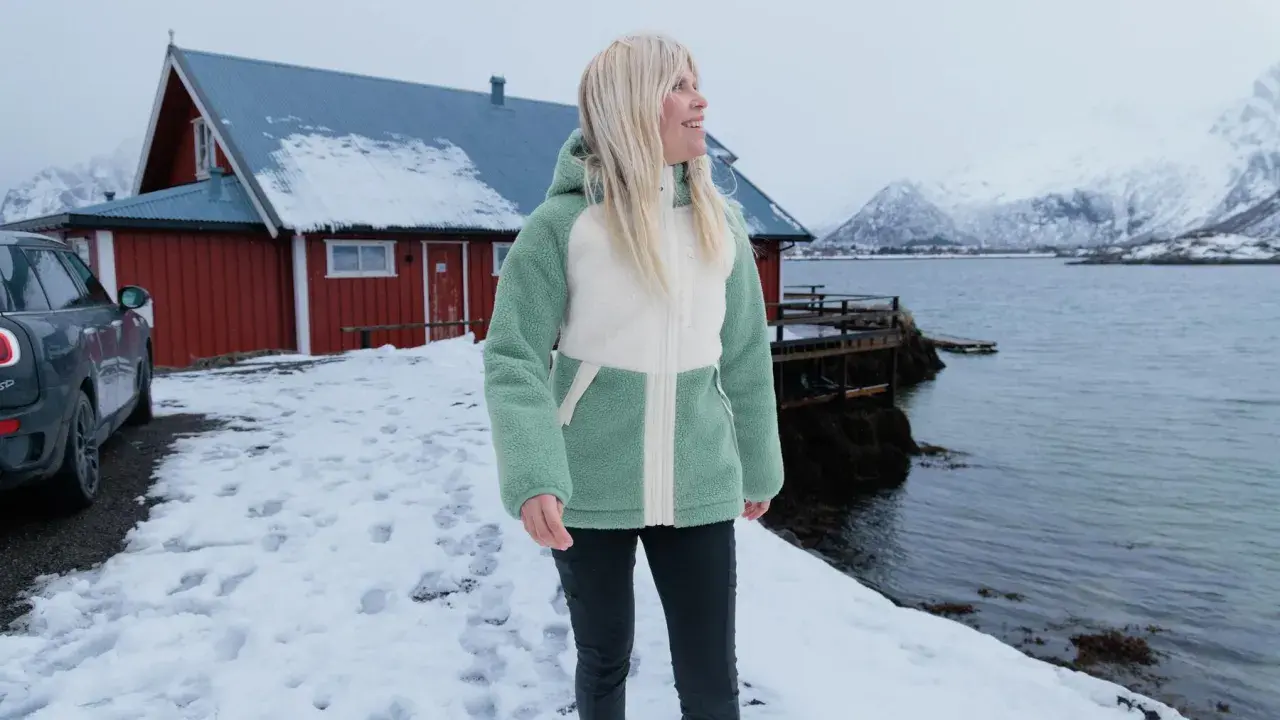 Frau in flauschiger Fleecejacke, bereit für das **fleece waschen**, steht vor rotem Haus am Meer.