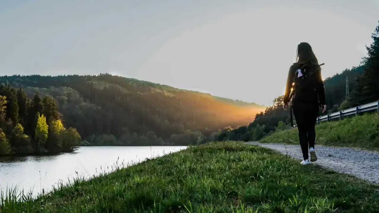 Frau mit Rucksack wandert auf einem Weg am See im Odenwald. Sonnenuntergang beleuchtet die Hügel.