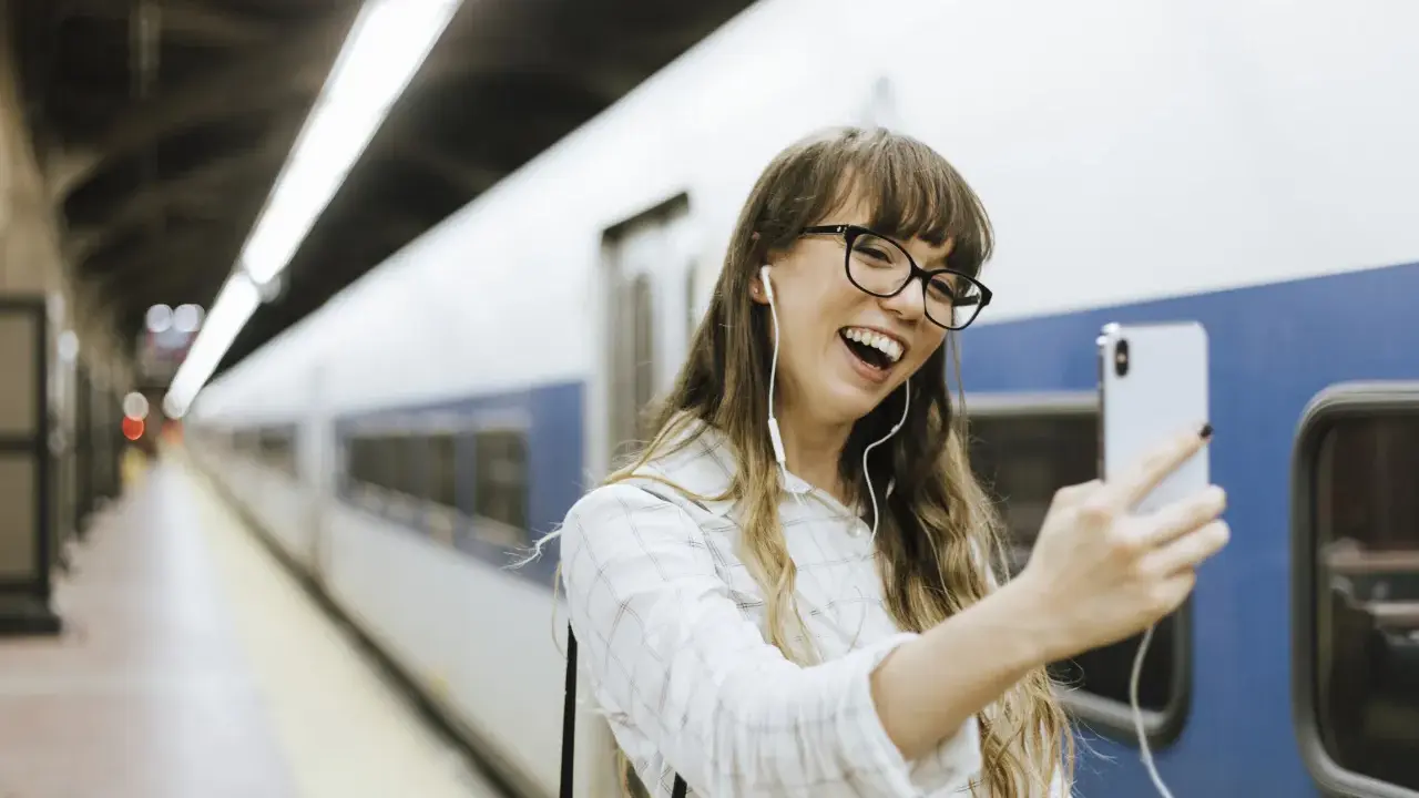 Junge Frau mit Kopfhörern macht ein Selfie am Bahnhof. Sie freut sich, ihr nächstes Abenteuer mit dem **discover eu pass** zu beginnen.