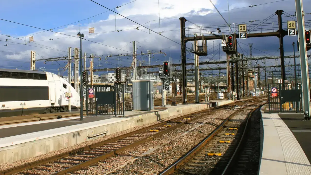 Ein weißer Hochgeschwindigkeitszug steht am Bahnsteig. Die Gleise führen in die Ferne, vorbei an Signalen und Oberleitungen. Vielleicht ist dies der **Zug nach Barcelona**?