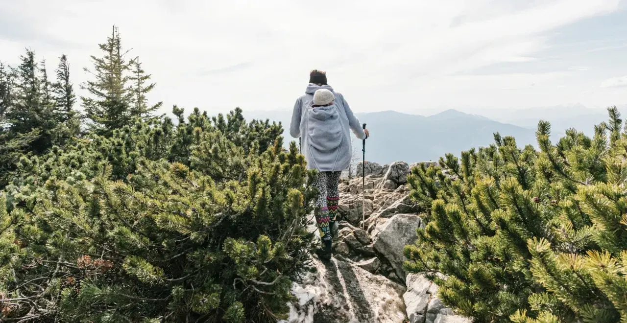 Frau wandert mit Baby im Tragetuch auf einem felsigen Bergpfad, umgeben von grünen Kiefern.