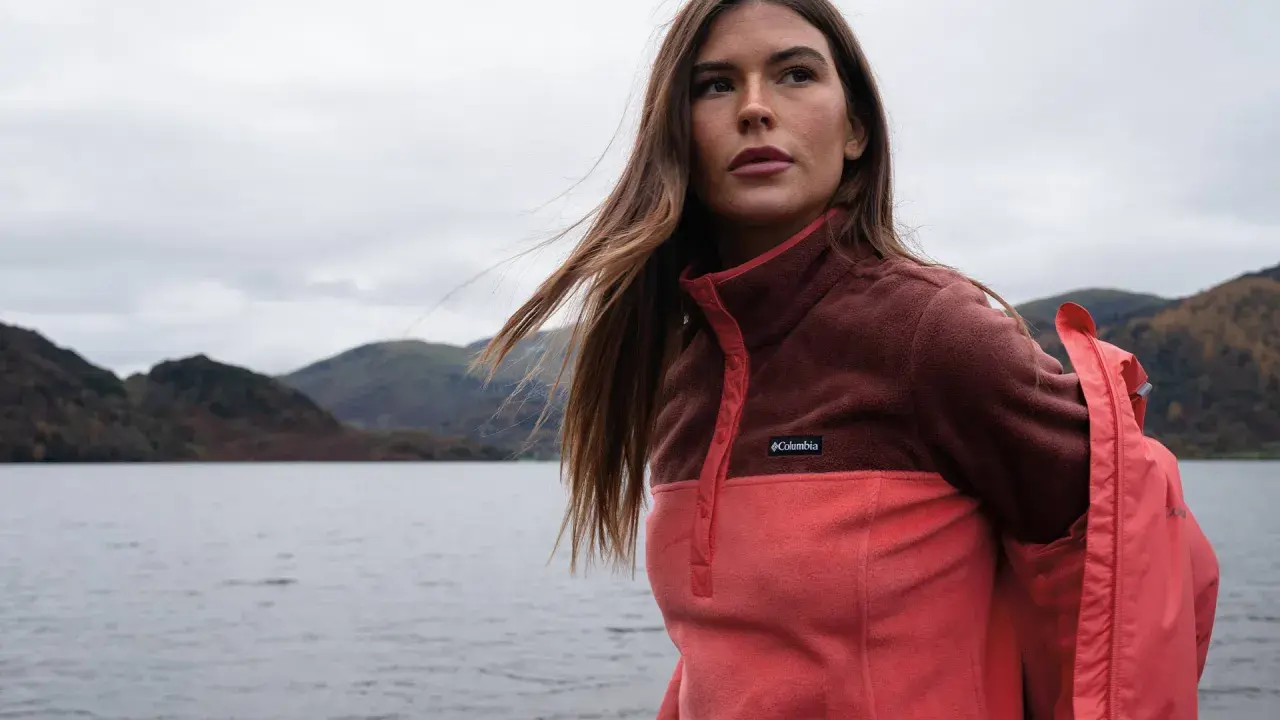 Frau mit langen Haaren im Zwiebel-Look, trägt eine zweifarbige Fleecejacke von Columbia am Seeufer.