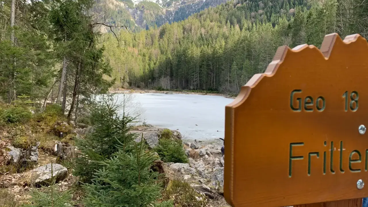 Holzschild "Geo 18 Fritter" vor gefrorenem See und Bergkulisse. Perfekt für einen Sommerurlaub in Bayern.