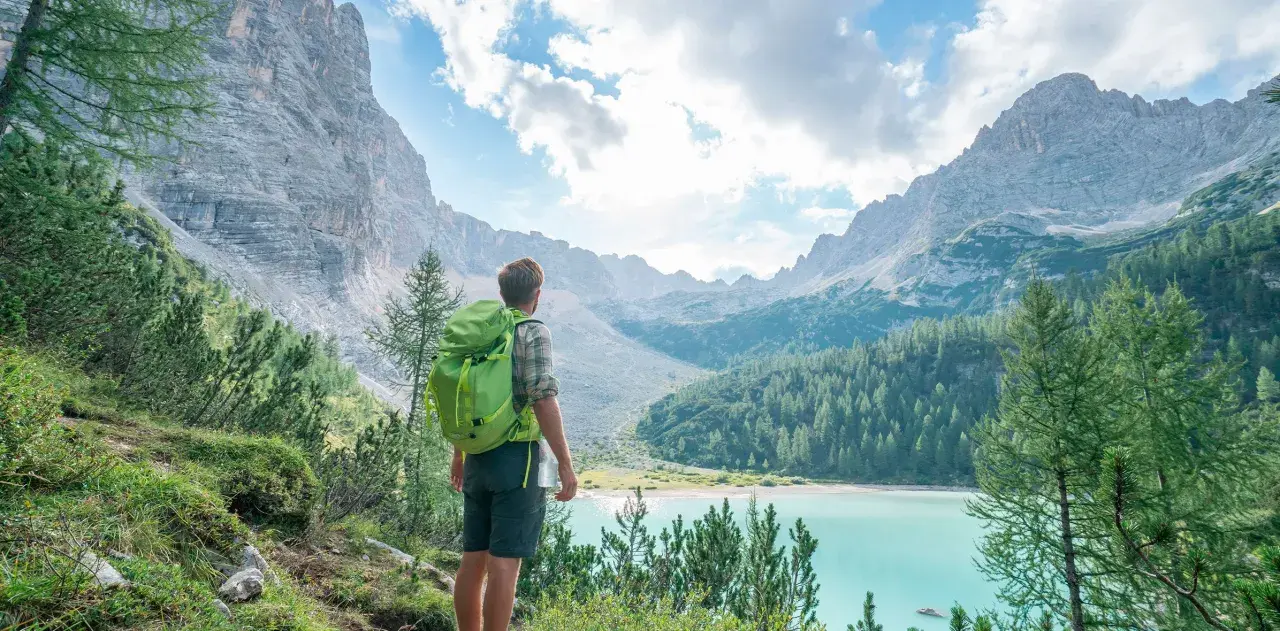 Wanderer mit Rucksack blickt auf türkisfarbenen Bergsee. Die Alpenlandschaft erinnert an die Grenze zwischen Schweiz und Italien.