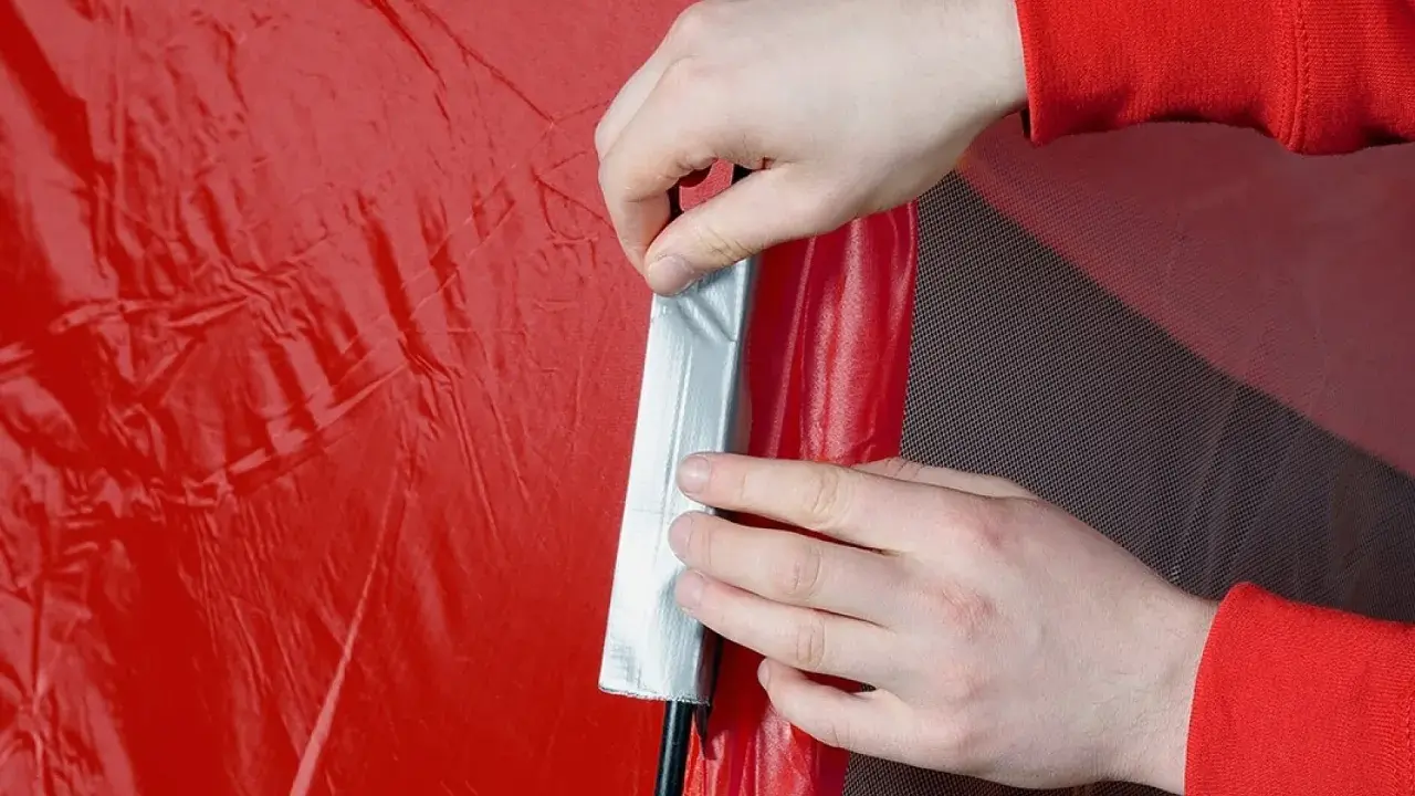 Hände reparieren ein rotes Zelt mit Klebeband.