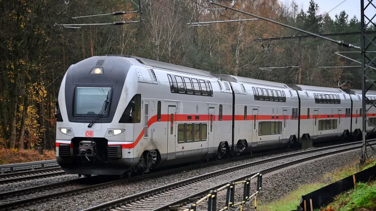 Ein moderner Doppelstockzug der Deutschen Bahn fährt durch eine herbstliche Landschaft. Die roten Streifen am Zug heben sich vom silbernen Lack ab.