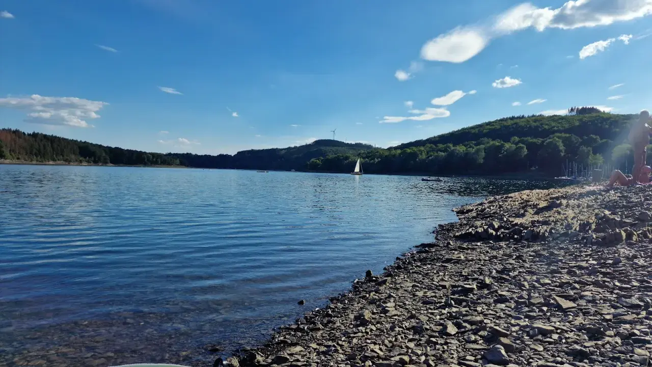 Sonniger Tag am See in NRW. Ein Segelboot gleitet über das Wasser, während am steinigen Ufer Menschen entspannen.