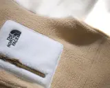 Ein beige-weißes Fleece-Oberteil von The North Face mit Reißverschlusstasche. Fleece ist ein weiches, warmes Material.