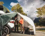 Zwei Frauen genießen das Frauen Camping Abenteuer am See. Ein Zelt ist am Auto befestigt, mit Blick auf Berge und Wasser.