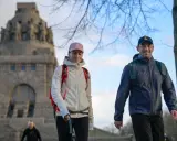 Ein Paar genießt eine Wanderung, passend mit wetterfester Wanderkleidung und Rucksäcken ausgestattet, vor einem imposanten Denkmal.