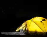 Gelbes Zelt leuchtet in der Nacht unter Sternenhimmel. Stiefel stehen bereit. Alles, was man zum Zelten braucht, ist hier.