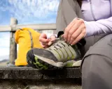 Person bindet Schnürsenkel an schmutzigen Wanderschuhen. Gelber Rucksack und Holzplattform im Hintergrund.