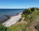Ein sonniger Tag am Timmendorfer Strand. Die Ostsee glitzert, ein schmaler Kiesstrand lädt zum Spaziergang ein, während grüne Büsche die Steilküste säumen.