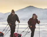 Zwei Personen ziehen Schlitten durch verschneite Landschaft. Die Berge im Hintergrund sind mit Schnee bedeckt. Ein Abenteuer wie aus dem Fjällräven g-1000 Katalog.