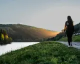 Frau mit Rucksack wandert auf einem Weg am See im Odenwald. Sonnenuntergang beleuchtet die Hügel.