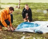 Ein Mann in einem orangen Anorak und eine Frau im Hintergrund bauen ein Zelt auf. Sie bereiten sich auf das **Zelt reinigen** nach dem Camping vor.