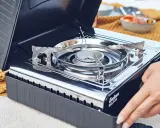 Tragbarer Gaskocher mit offenem Deckel, bereit für die Zubereitung von Speisen. Die Gaskartuschen lagern sicher im Inneren.