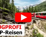 Ein roter Panoramazug fährt über eine Steinbrücke durch eine Berglandschaft mit Wäldern und schneebedeckten Gipfeln.