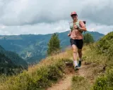 Lächelnde Frau beim Trailrunning Training auf einem Bergpfad, mit Blick auf grüne Täler und bewölkten Himmel.