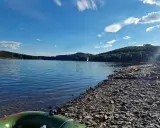 Sonniger Tag am See in NRW. Ein Segelboot gleitet über das Wasser, während am steinigen Ufer Menschen entspannen.
