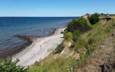 Ein sonniger Tag am Timmendorfer Strand. Die Ostsee glitzert, ein schmaler Kiesstrand lädt zum Spaziergang ein, während grüne Büsche die Steilküste säumen.