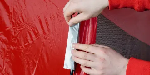 Hände reparieren ein rotes Zelt mit Klebeband.