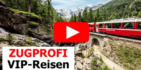 Ein roter Panoramazug fährt über eine Steinbrücke durch eine Berglandschaft mit Wäldern und schneebedeckten Gipfeln.