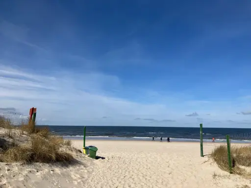 Ein sonniger Tag am Strand von Misdroy, Polen. Menschen spazieren am Meer entlang, während sich im Vordergrund Dünen und Strandgras erstrecken.