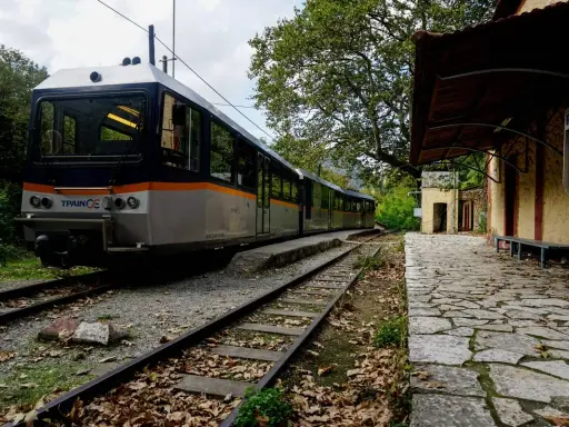 Ein Zug nach Griechenland steht auf den Gleisen neben einem alten Bahnhofsgebäude. Herbstlaub bedeckt den Bahnsteig.