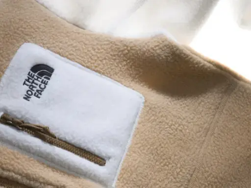 Ein beige-weißes Fleece-Oberteil von The North Face mit Reißverschlusstasche. Fleece ist ein weiches, warmes Material.