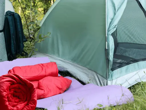 Zwei rote Schlafsäcke liegen auf lila Isomatten vor einem grünen Zelt. Ein Rucksack und eine weitere Isomatte stehen daneben.