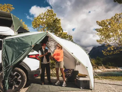 Zwei Frauen genießen das Frauen Camping Abenteuer am See. Ein Zelt ist am Auto befestigt, mit Blick auf Berge und Wasser.