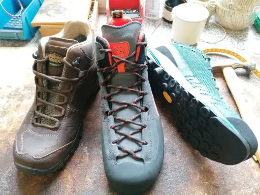 Drei Wanderschuhe von verschiedenen Herstellern: braun, grau und türkis. Werkzeuge und Garn liegen daneben.