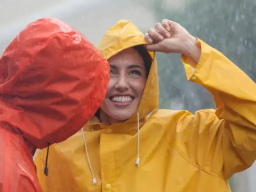 Zwei lachende Personen im Regen, eine in Gelb, die andere in Rot. Die gelbe Regenjacke ist im Fokus.
