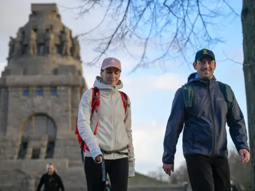 Ein Paar genießt eine Wanderung, passend mit wetterfester Wanderkleidung und Rucksäcken ausgestattet, vor einem imposanten Denkmal.