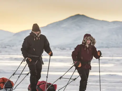 Zwei Personen ziehen Schlitten durch verschneite Landschaft. Die Berge im Hintergrund sind mit Schnee bedeckt. Ein Abenteuer wie aus dem Fjällräven g-1000 Katalog.