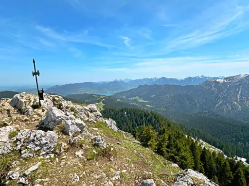 Gipfelkreuz am Fockenstein bei Lenggries, mit Blick über Wälder und Berge bis zum See.
