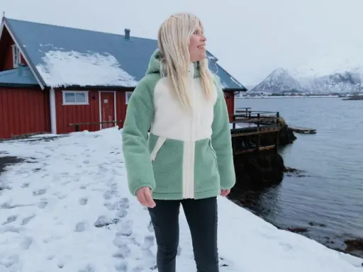 Frau in flauschiger Fleecejacke, bereit für das **fleece waschen**, steht vor rotem Haus am Meer.